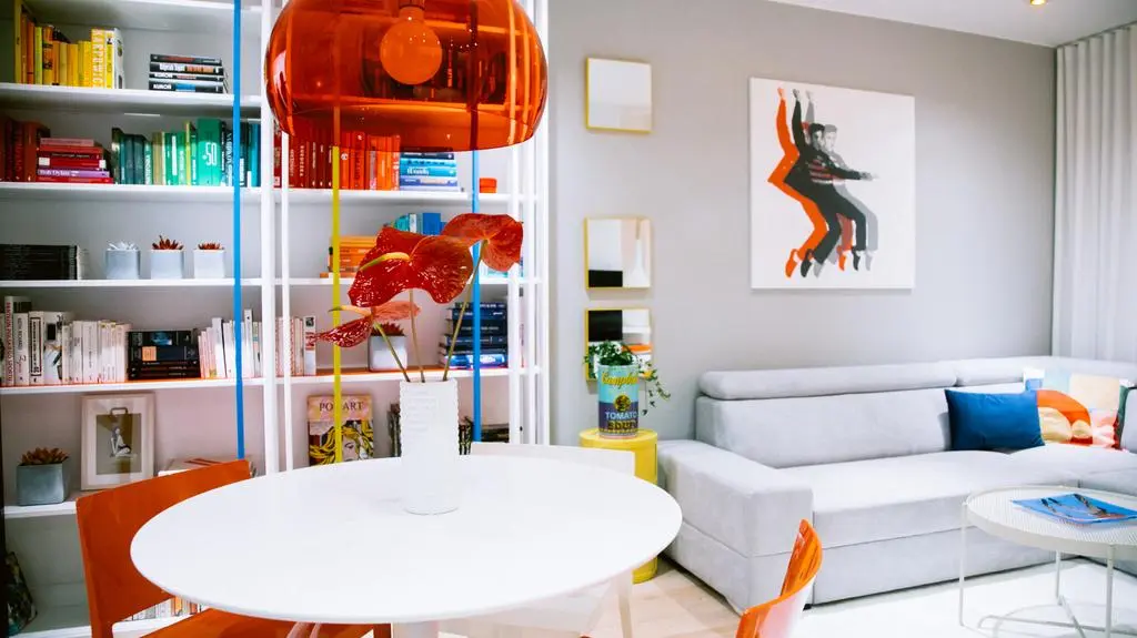 Szara kanapa na tle szarej ściany oraz meble stały się bazą do ekspozycji kolorowych dodatków, nadających wnętrzu charakteru i nawiązujących do stylu pop-art.