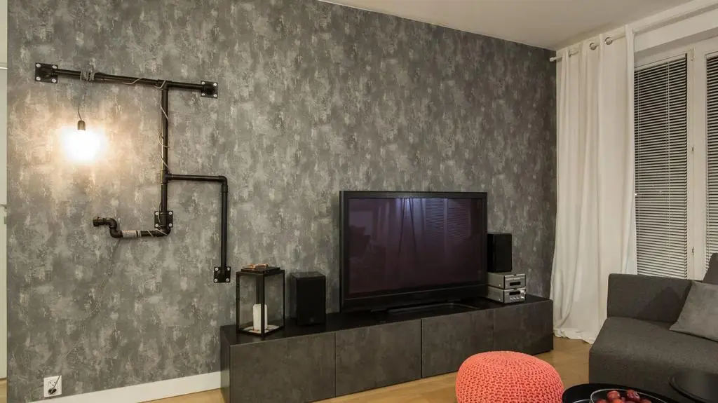 Największą metamorfozę w salonie przeszła ściana przylegająca do sypialni. Położono na niej tapetę imitującą beton i zamontowano na niej lampę z rur. Wymieniono też szafkę pod telewizorem.