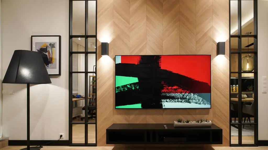 Lustra w loftowych ramkach nawiązujących do ścianki wydzielającej część do pracy oraz nowoczesny telewizor umożliwiający wyświetlanie ulubionych grafik lub zdjęć świetnie uzupełniają salon.